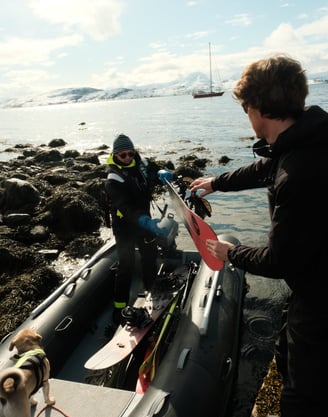 Two skiers unloading gear from a dinghy with snowy Lyngen Alps and a sailboat in the background