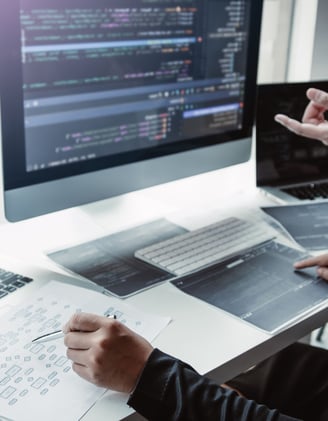 two people collaborating at a desk with laptops, a desktop and printouts