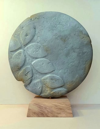 A round plate-like sculpture depicting leaf-shaped motifs, painted in blue greys, on oak plinth.