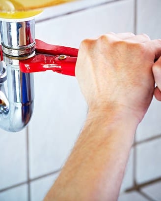 man using a red mini pipe wrench to turn a pipe under the sink