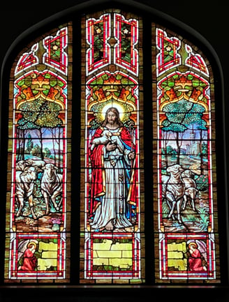 A stained glass window featuring Jesus and several sheep, Jesus holds a lamb in his arms