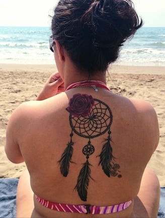 Mulher de costas na praia, com tatuagem de rosa e filtro dos sonhos, símbolo de memória ancestral