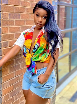 Efia Odo in a designed shirt and jeans shorts standing close to a wall.