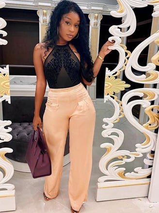 Efia Odo poses in a black net top, in a cream trousers and a mauve handbag.