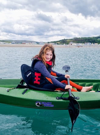 A girl kayaking in a sit on top kayak in the sea with Borth village and cloudy skies behind her 