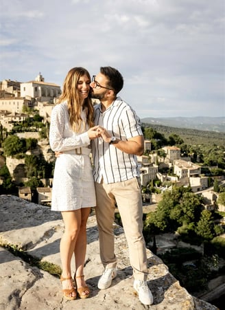 Editorial engagement photo of man kissing fiancée on cheek with scenic view of Gourdon village