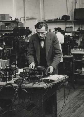 John Logie Baird in a tweed suit and bow tie adjusts a rotating Nipkow disc assembly
