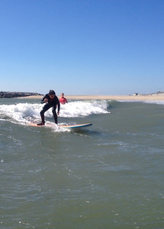 A young male surfer rides an unbroken wave with sunny skies while a surf instructor looks on