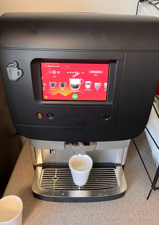 Super-automatic coffee machine dispensing a cappuccino into a paper cup with touchscreen interface.