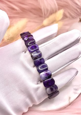A gloved hand holding a deep purple beaded bracelet against a soft, neutral-toned background.