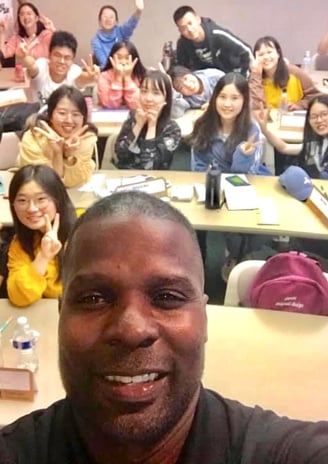 Dr. Marlon Ware standing in front of his class with students holding up the peace sign