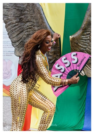 Eine Drag Queen posiert in einem goldenen Overall mit einem rosa Fächer.