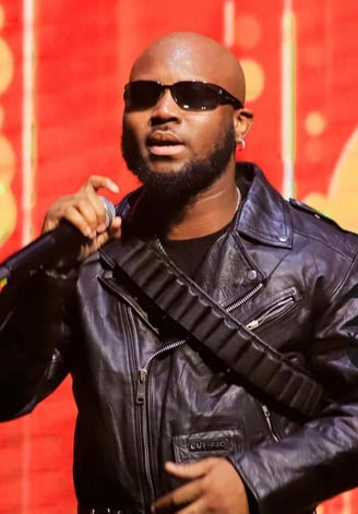 King Promise in a black vest holding a microphone in dark spectacles.