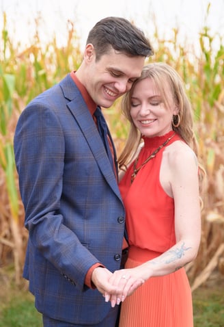 a man in a blue suit and woman in an orange dress holding hands in front of a corn field