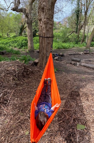 Child lying in an orange hammock in a woodland.