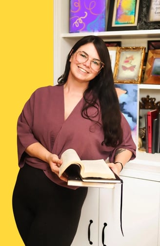 A woman stands leaning on a bookshelf with two books in her hand, one is open