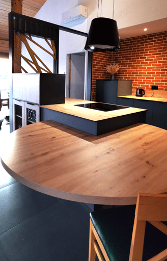 Cuisine vert foncée avec table en bois centrale posée sur îlot avec hotte suspendue. Plan compact