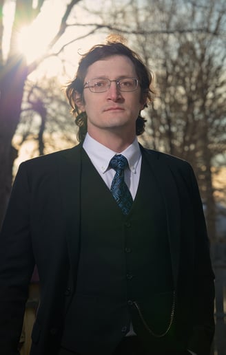 a man in a  black suit and glasses and tie with a blue tie in front of the sun and trees