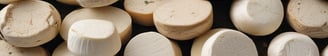 Two large, rustic wheels of cheese, one partially cut, are stacked on top of each other against a rough stone background. The cheese has a natural, textured rind and the cut reveals a smooth, pale interior.
