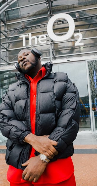 Medikal posed in a black vest with headphones in front of the O2 Arena.