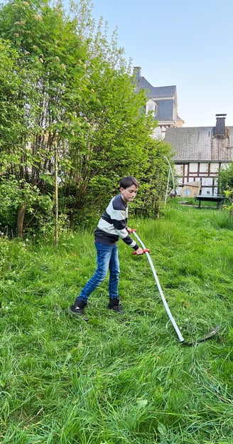 Philipp Geisler mäht mit einer Sense die Wiese in seinem Garten