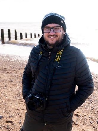 a profile photo of Ben Walker with his Nikon D500 hanging from his neck. Wearing a black & grey beanie standing on the beach