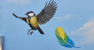 Fresque peinte sur une façade. Une mésange et une plume colorée s'envolent sur un ciel bleu.