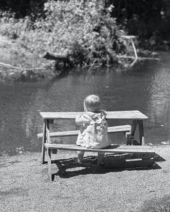 Black and white photo of a baby girl sitting at a miniture picnic table, gazing at a lake with trees in the distance.
