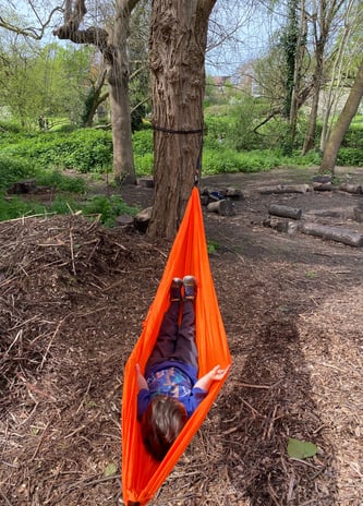 Child lying in an orange hammock in a woodland.