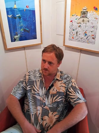 Artist seated in a red chair beneath framed digital prints of underwater and courthouse-themed artwo