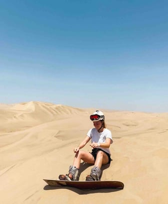 Manon sitting in the sand, ready to ride down the dunes of Huacachina on a sandboard, in the Peruvian desert.