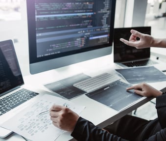 two people collaborating at a desk with laptops, a desktop and printouts