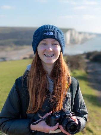 a profile photo of Lilie Walker, smiling holding a Nikon D5300 against a backdrop of Cuckmere Haven and Seven Sisters