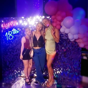 3 girls posing for a photo in front of a silver shimmer backdrop and balloon garland with Happy 18th neon sign hire Brisbane.