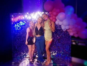 3 girls posing for a photo in front of a silver shimmer backdrop and balloon garland with Happy 18th neon sign hire Brisbane.