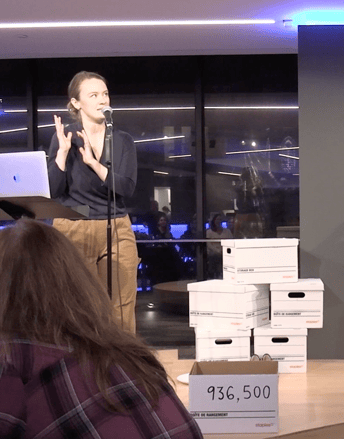 A white woman speaks at a microphone beside a pile of white fileboxes