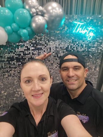 Carly and Kahn from SweetAz Co. taking a selfie in front of the silver shimmer wall, Thirty neon sign, and balloon garland fo