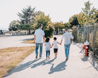 Two dads walking and holding hands with their children on a wide sidewalk in the sun