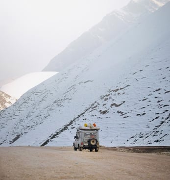 An old style land Rover on a dirt road beside a snow covered mountain
