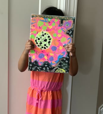 little girl with dark hair in a pink and orange dress holds colorful painting in front of her face