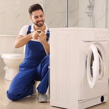 A smiling male technician is crouching down to repair the side panel of a white dryer with a screwdr
