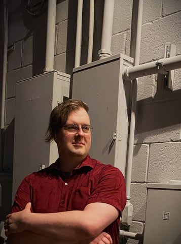 photo of G.J., a 40-ish white person, arms folded, standing before an industrial circuit breaker at night.