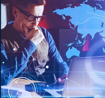 Business leader in glasses works on a laptop at a desk, one hand over his mouth, with a holographic continent behind him.