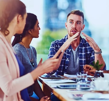 A man sits at a table, using hand gestures to explain something to four others seated around him.