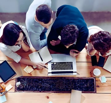 A group of leaders huddle around a laptop at a table, discussing the content on the screen.