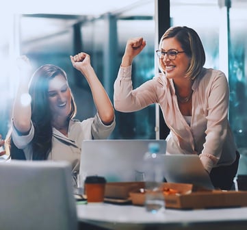 Group of leaders collaborates at a boardroom table, focusing on two female leaders celebrating their win with fist pumps