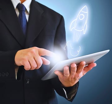 A man in a black suit and tie holds a tablet, touching the screen with his index finger as a holographic rocket ship emerges.