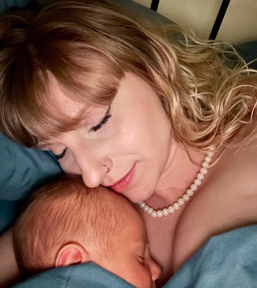 A mother with blonde hair and a pearl necklace peacefully cuddles her sleeping newborn baby, both wrapped in a blue blanket