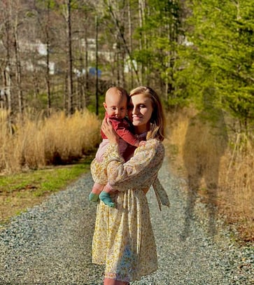 A mother holding baby on a gravel road at dusk, with the shadow of the father looming behind—tension of co-parenting & abuse 
