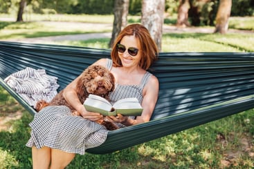 A woman relaxing in a hammock with her dog, reading a book outdoors, symbolising stress management.